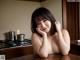 A naked woman sitting at a kitchen counter with her hand on her chin.